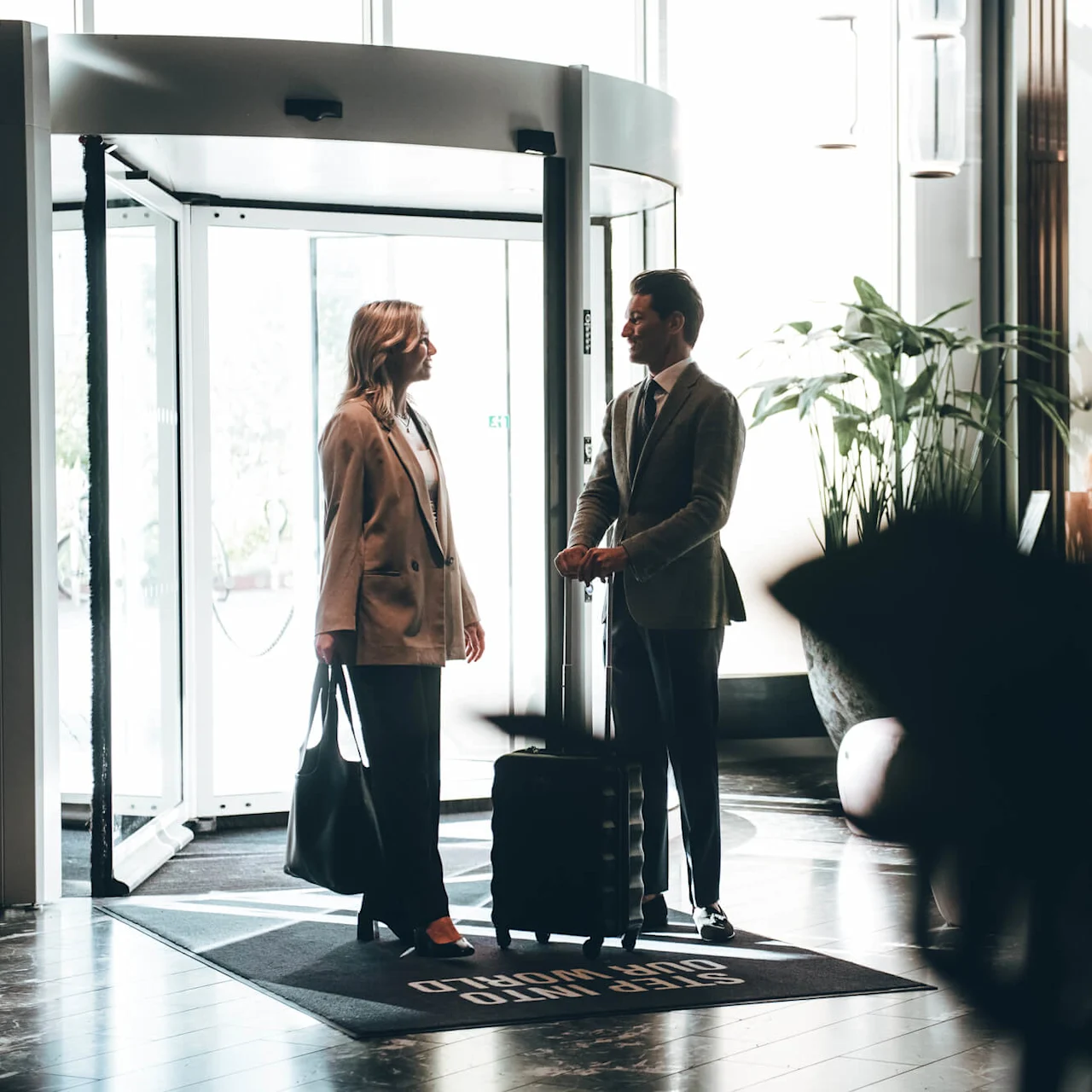 Two people, one with a suitcase, converse casually in a modern hotel lobby. Bright lighting from large windows highlights decor, including a large plant. Text on mat: "STEP INTO OUR WORLD."