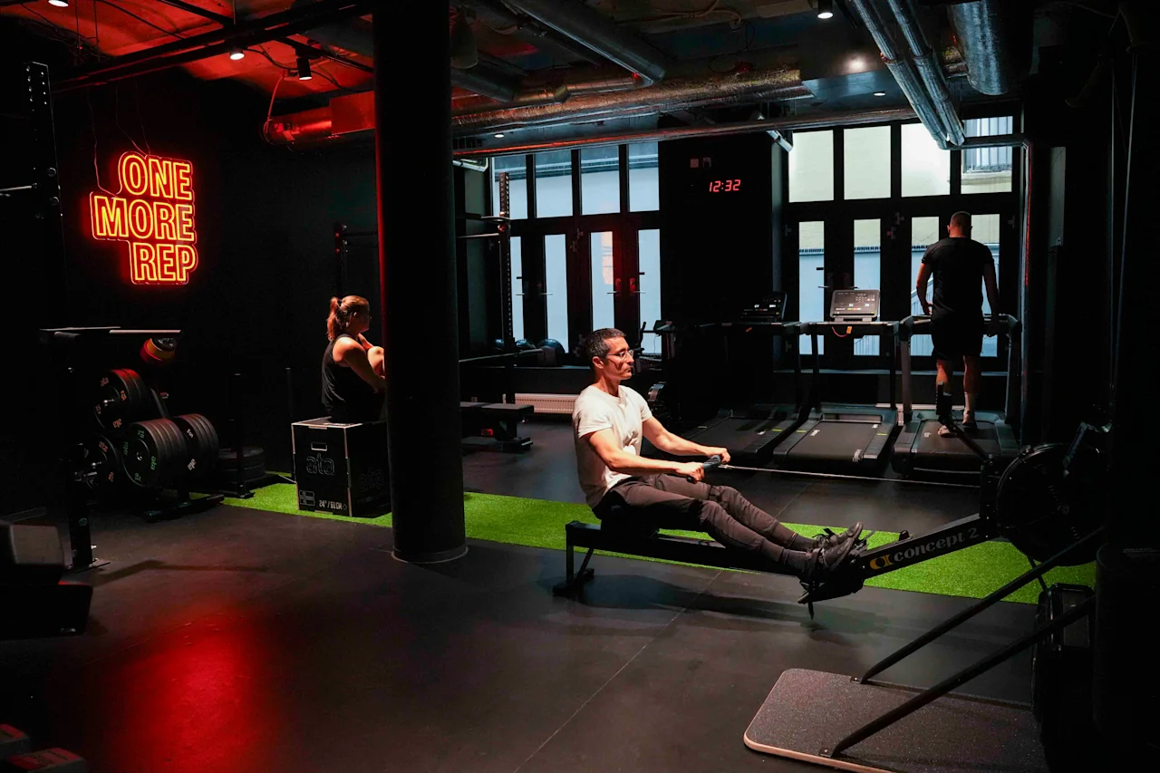 En person ror på en romaskine i et fitnesscenter, under et neonskilt med teksten "ONE MORE REP". To personer anvender andet træningsudstyr i baggrunden, under dæmpet belysning.