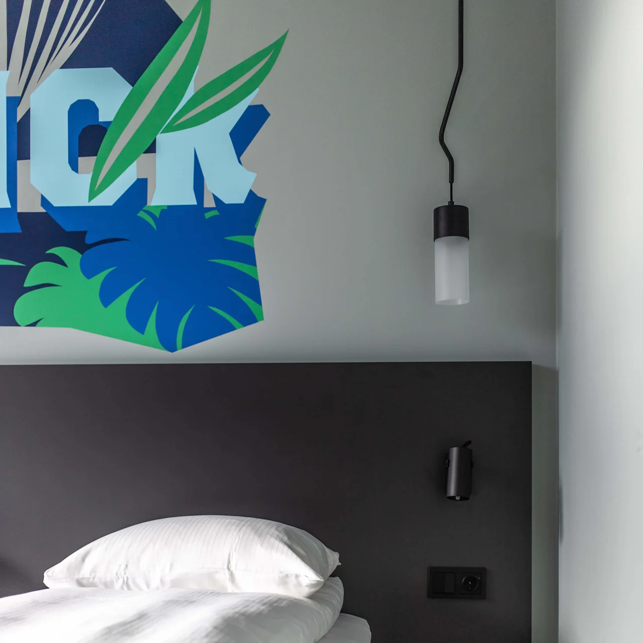 A bed with white sheets sits against a dark headboard; a pendant light hangs above. Wall decor shows the text "ICK" with green leaves.