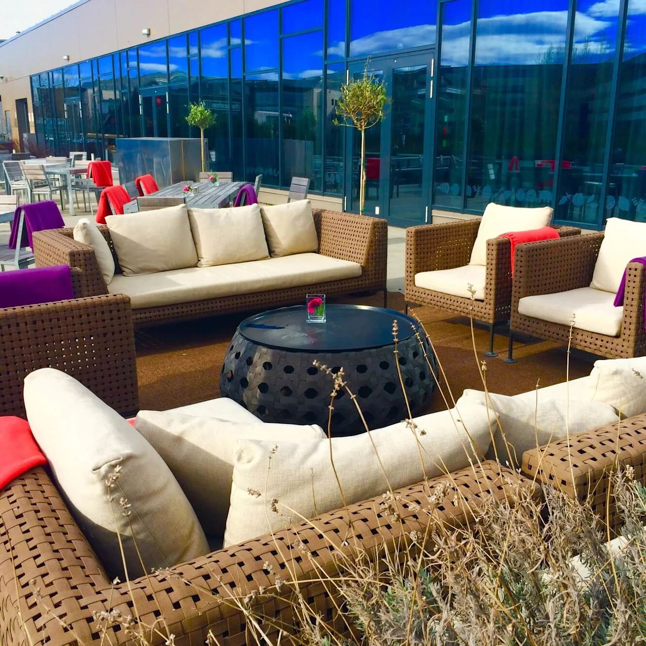Wicker patio furniture with beige cushions surrounds a black metal table under clear skies. Red and purple cloths drape chairs next to glass-walled building reflecting clouds.