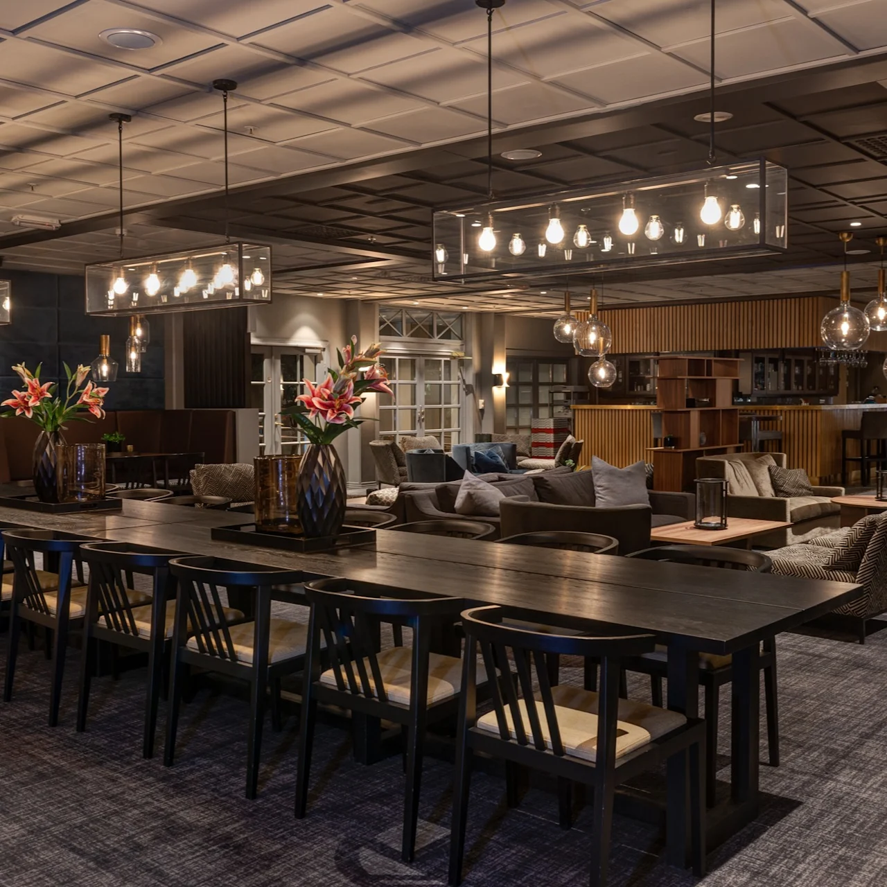 Large table with black chairs hosts vases of pink flowers, under modern pendant lights in an elegant lounge featuring armchairs, bar, and softly lit ambiance.
