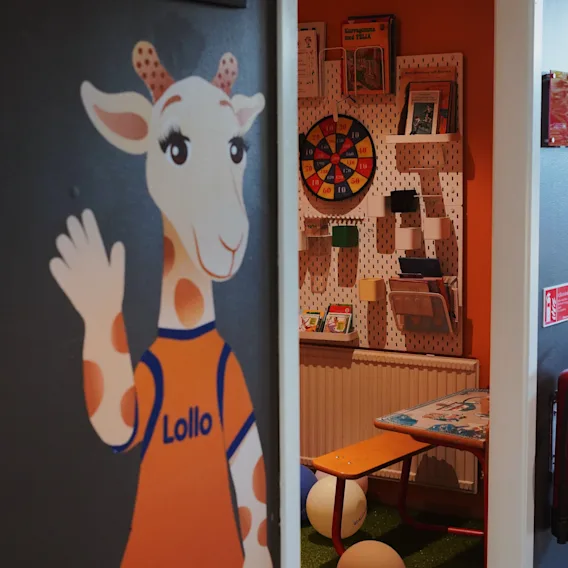 Tecknad giraff vinkar bredvid en dörröppning som leder till en färgglad lekplats för barn med darttavla och litet bord.