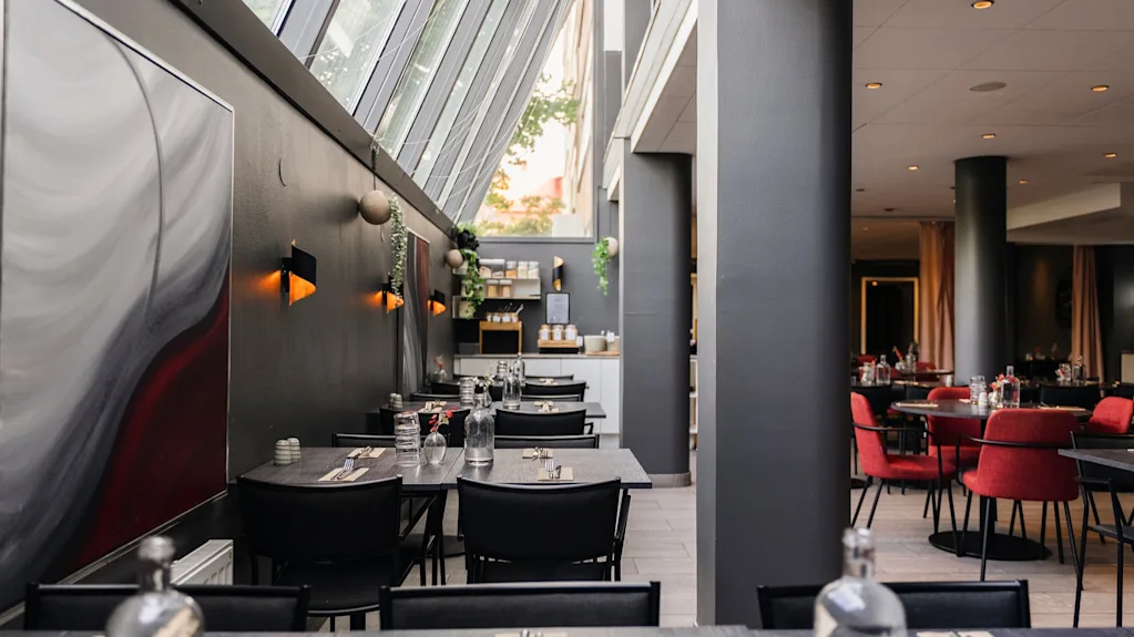 Samtida restauranginteriör med prydligt dukade bord med svarta stolar, grå väggar prydda med abstrakt konst och ett stort snedställt fönster som släpper in naturligt ljus. Röda stolar fyller den rymliga matsalen.