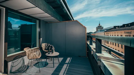 To rottingstoler og et lite rundt bord står på en moderne takterrasse under en klar blå himmel. Smale bygninger og et fjernt tårn er synlige i horisonten.