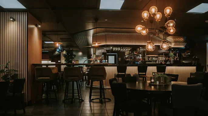 Et dunkelt belyst bar med krakker, bord og taklamper; ingen gjester til stede, rolig atmosfære.