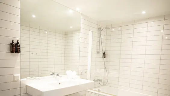 A white sink with a chrome faucet is mounted in a modern bathroom. It is reflected in a large mirror above, adjacent to a bathtub with a showerhead.