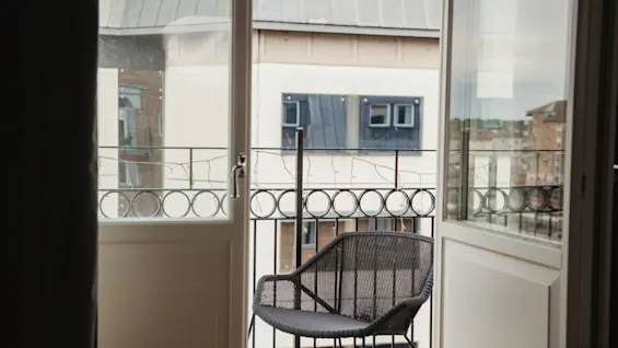 A black metal chair sits quietly on a small balcony. Open doors reveal city buildings with windows opposite, creating an urban atmosphere.