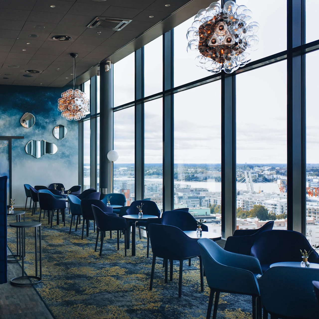 Stole og borde linjer en moderne lounge med blå vægge, dekorative spejle og kunstneriske loftlamper, ved siden af et panoramavindue med udsigt over et bylandskab. Høje planter forbedrer stemningen.