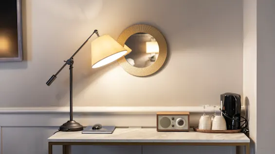 A well-lit hotel room desk at Home Hotel Grand Bodø with a lamp, mirror, radio, and coffee station, creating a cozy atmosphere.