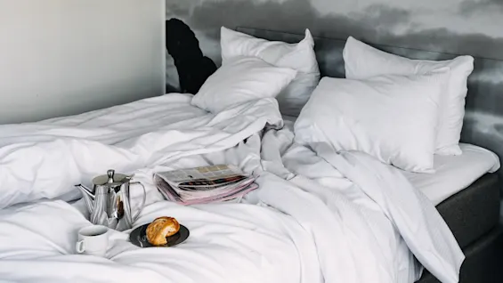 Bed with rumpled white sheets, holding a silver teapot, a cup, a croissant on a plate, and a newspaper. Two pillows rest against a headboard with cloudy wall art.
