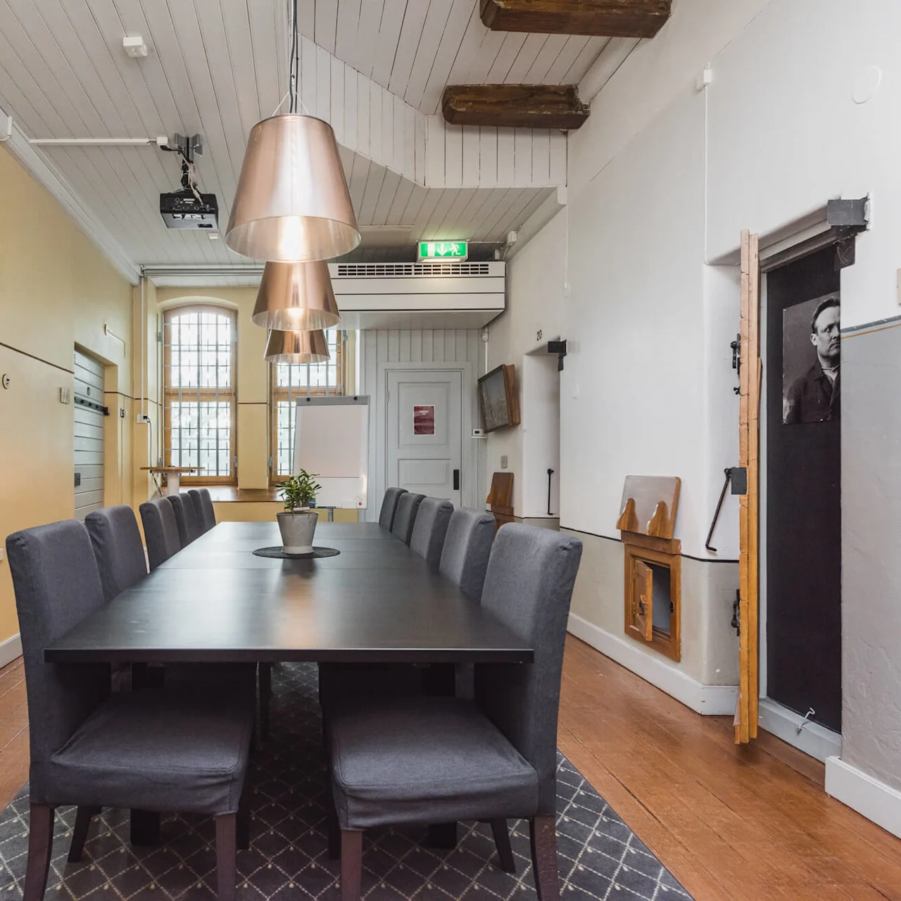A conference room with a large table, chairs, and a pendant lamp. Wooden floors and traditional windows suggest a historical setting.