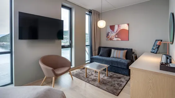 Et hyggeligt hotelværelse har en sofa og lænestol ved gulv-til-loft-vinduer. Et vægmonteret TV, et bord og et skrivebord med lampe fuldender den moderne indretning.