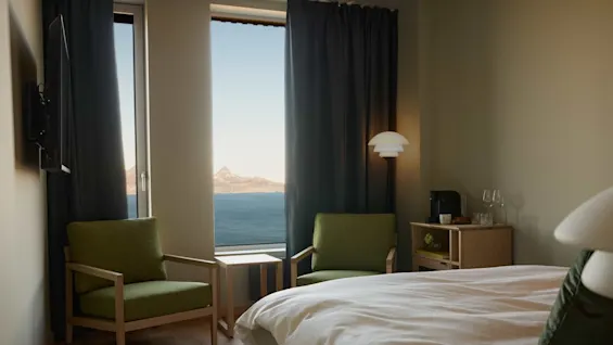 Room with two green armchairs facing a large window with a mountain and sea view; bed and side table with a coffee maker beside.  