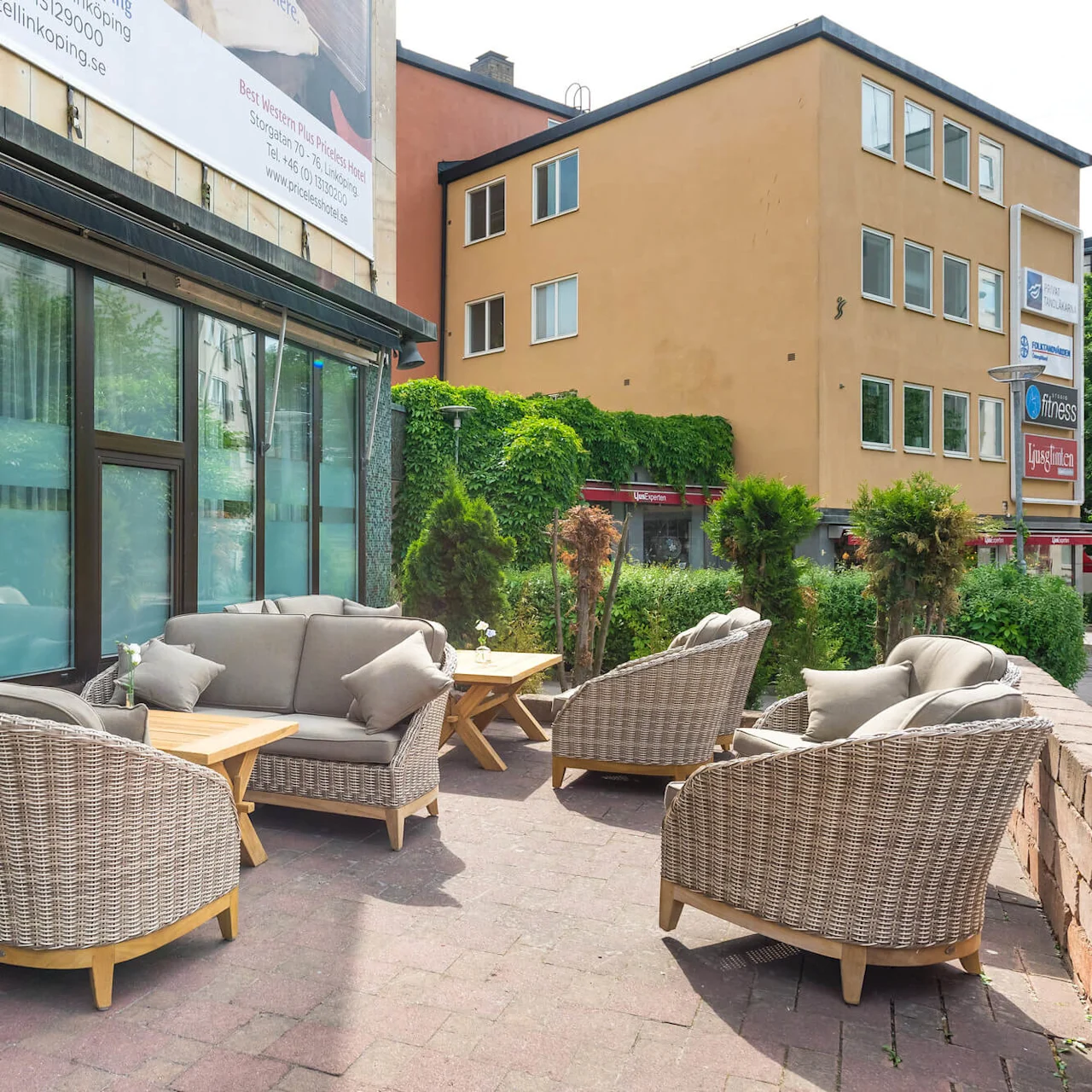 Rottingstolar och en soffa med kuddar är arrangerade på en stenlagd uteplats utanför en byggnad med stora fönster.