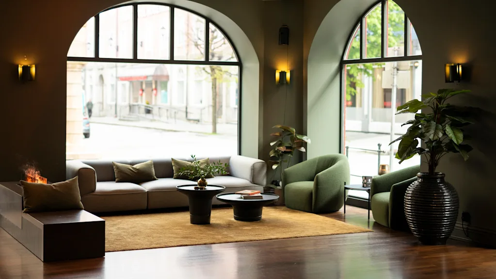 En modern lounge har en soffa och fåtöljer runt två små bord på en matta, upplyst av stora välvda fönster med gatutsikt. Krukväxter ger grönska.