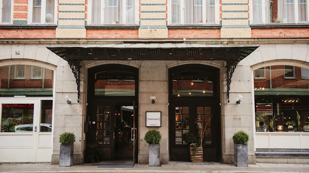 En charmerende bygningsindgang med to mørke døre, en udsmykket markise og potteplanter. Et skilt læser TEMPERANCE HOTEL.