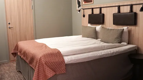 A neatly made bed with twin pillows and a rust-colored throw blanket stands against a wood-paneled wall with mounted lamps, beside a wooden door in a cozy, carpeted room.