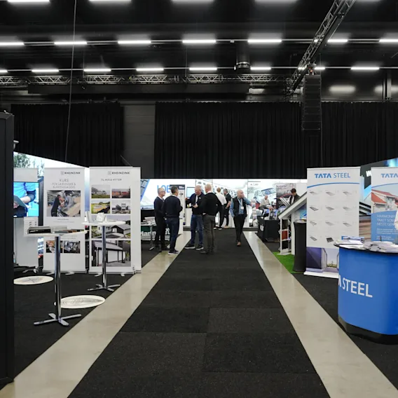 A trade show features exhibition booths with promotional displays from companies like Rheinzink and Tata Steel. Attendees walk and engage in conversations under bright lighting in a large hall.