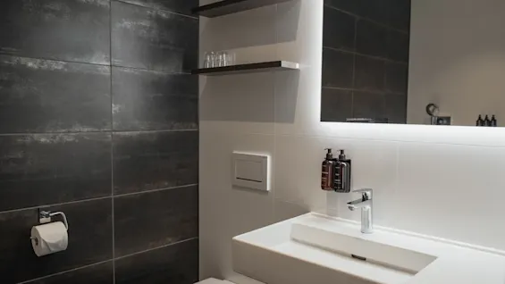 Et moderne badeværelse med en hvid vask, oplyst spejl og mørke flisevægge. Hylder med glas over toilettet.