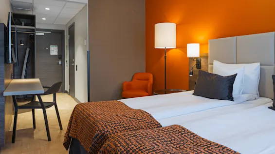 Ett hotellrum med en prydligt bäddad säng med orange och svart mönstrade överkast, en sänglampa, en orange accentvägg, en fåtölj och ett litet skrivbord.