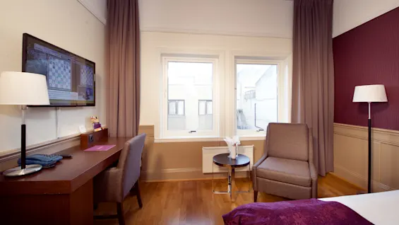 Et hotellrom interiør med en stor seng, et skrivebord med en fjernsyn, en stol, og vinduer som ser ut mot en bygning.