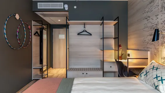 A modern hotel room with a neatly made bed, a wall-mounted shelving unit, and a work desk featuring a lamp. Three colorful hula hoops adorn the wall beside a door.