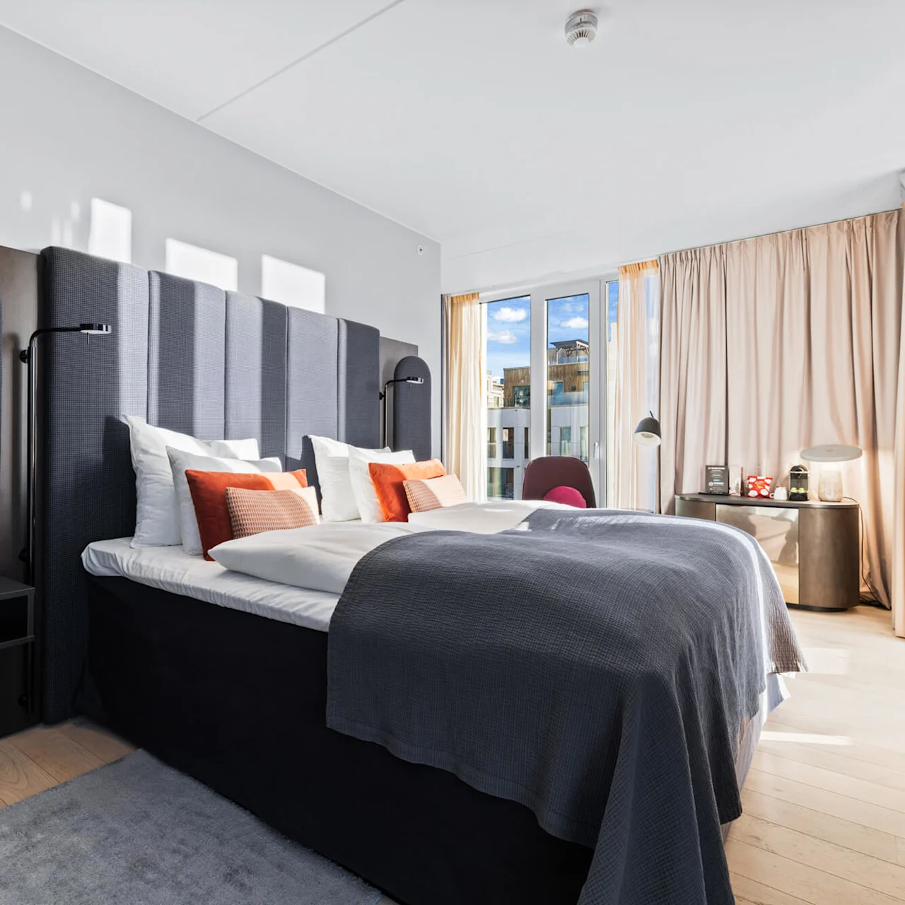 A large bed with gray and orange pillows stands in a modern, sunlit room. Floor-to-ceiling windows and light curtains create an airy, inviting atmosphere.