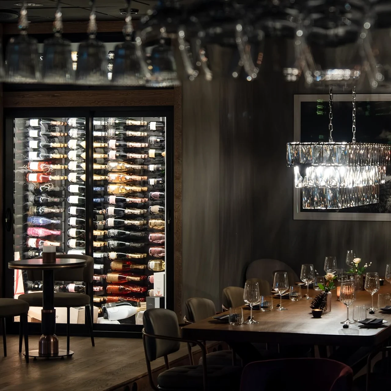 A dimly lit dining room features a long wooden table set with glassware and decorations, a large wine fridge filled with bottles, and a chandelier overhead.