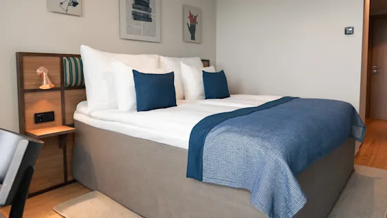 Et moderne hotellrom med en komfortabel seng, hvite sengetøy, blå puter og et matchende blått pledd.