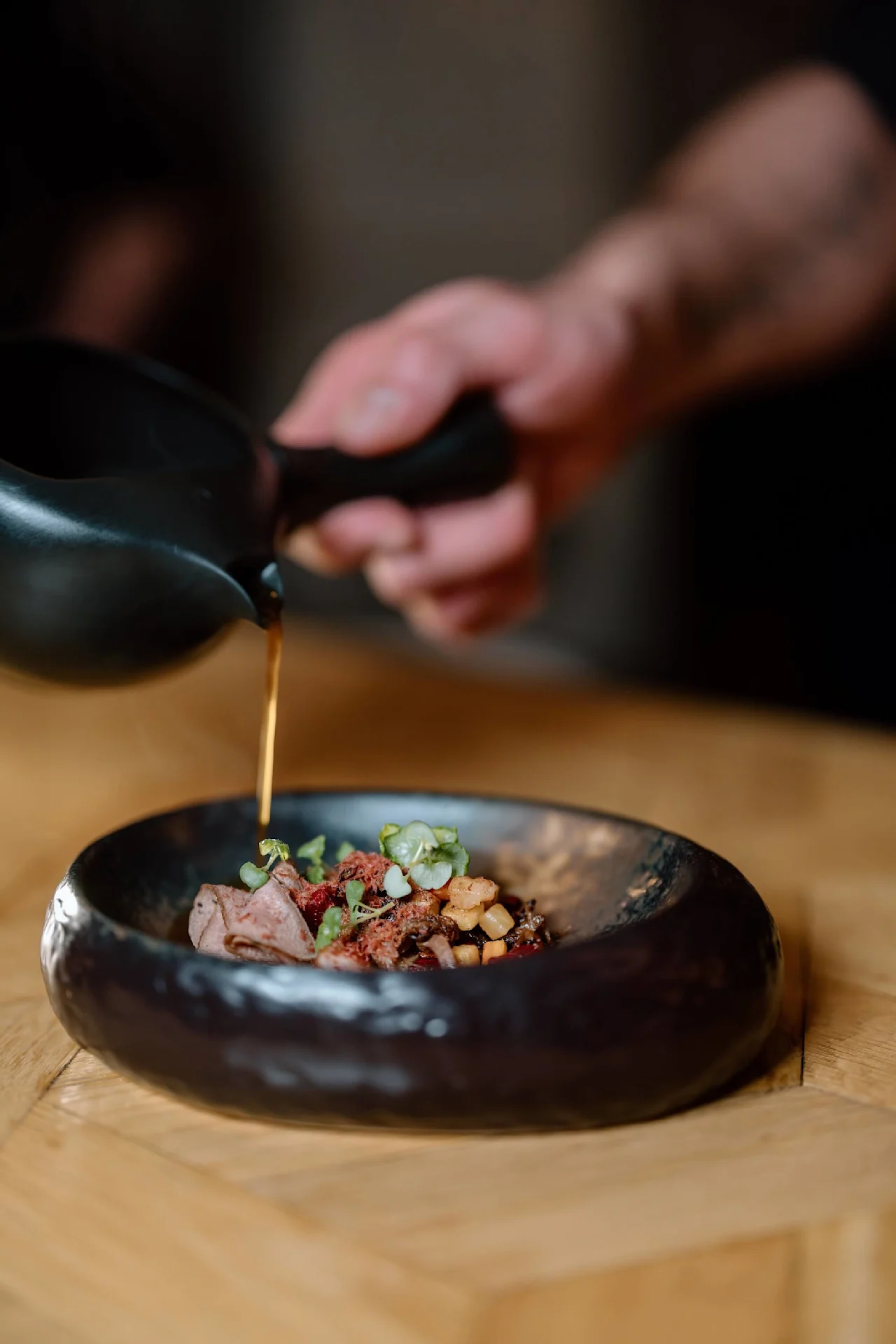 En mørk keramisk skål rummer en garneret ret, mens en hånd hælder sauce fra en sort kande. Miljøet er et træbord.
