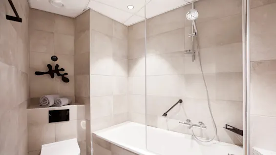 A modern bathroom with a white bathtub and shower fixture. Two rolled towels sit atop a shelf beside a toilet. Beige tiles cover the walls and floors.