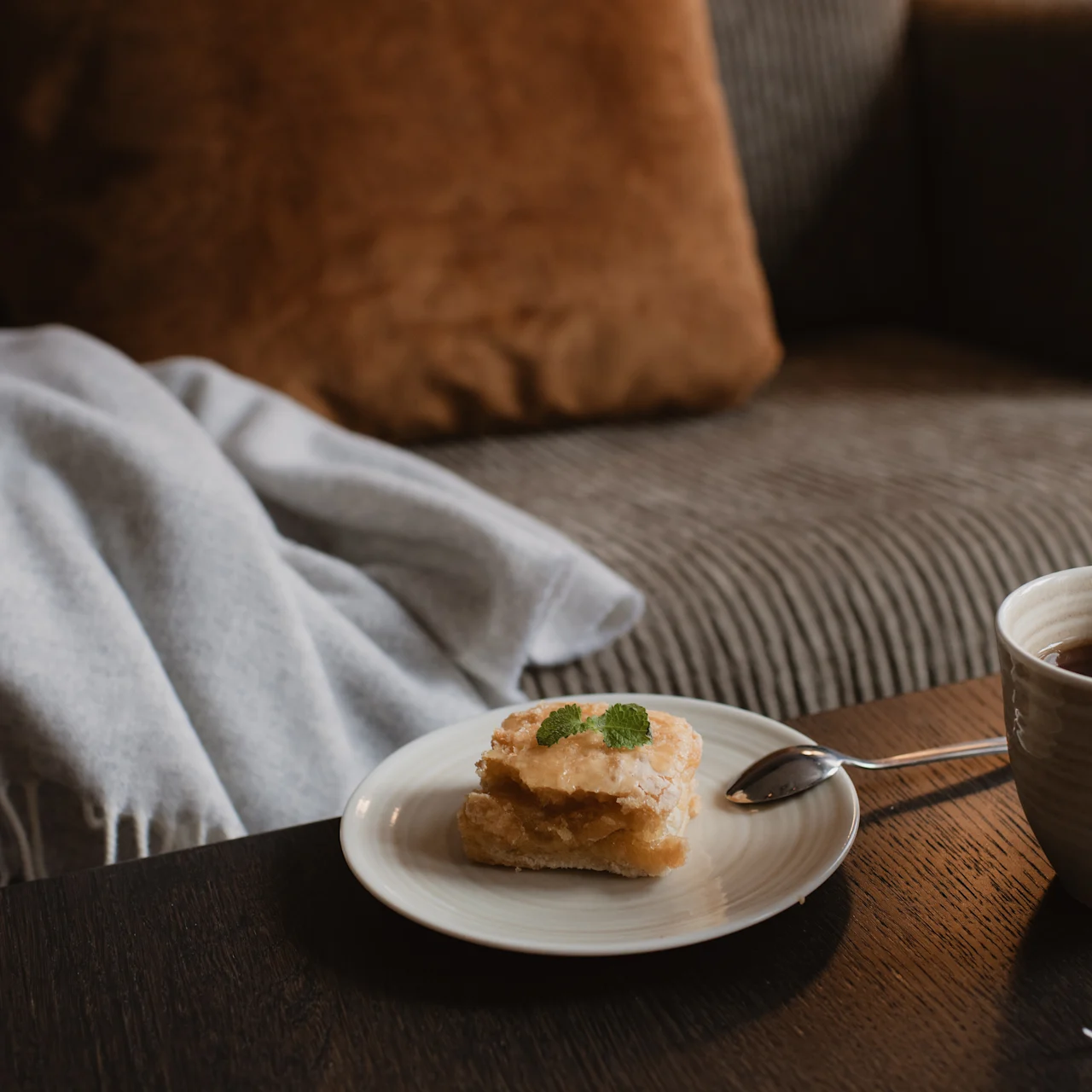 Pastry sits on a white plate with a spoon, next to a cup of tea on a wooden table. A cozy sofa and blanket provide a warm background.