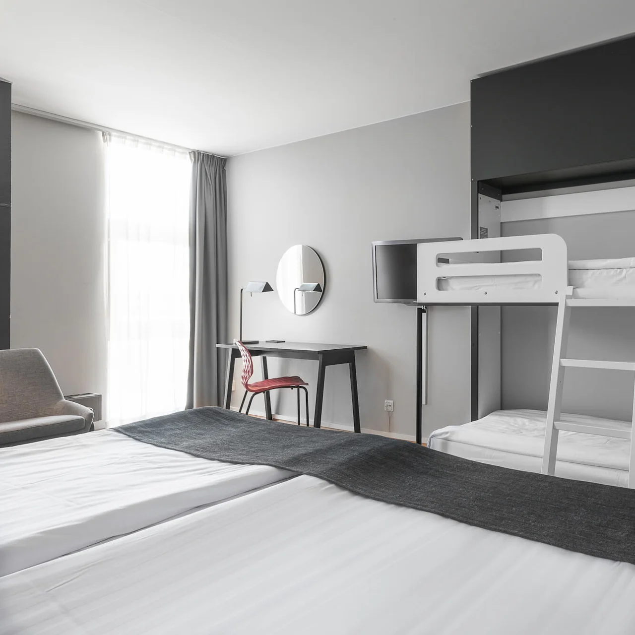 Bunk bed with white linens sits against a wall. A ladder connects the beds. A nearby work desk has a chair, lamp, and round mirror. A window illuminates the room.