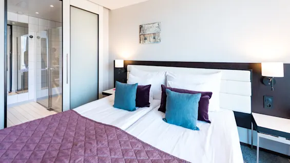 En pent oppredd seng med lilla og blå puter står i et moderne hotellrom, flankert av vegglamper og ved siden av en frostet glassdør til badet under en nøytral vegg.