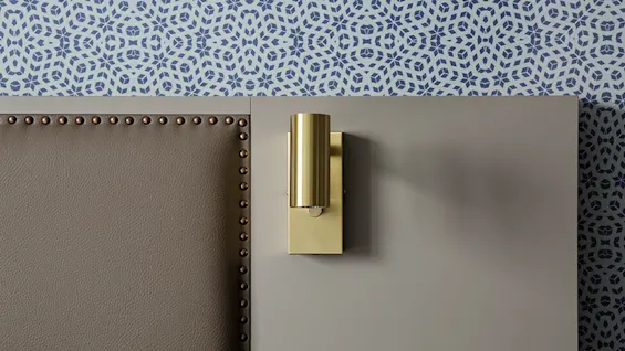 A gold wall sconce casts a shadow on a neutral wall above a studded couch, against patterned wallpaper.