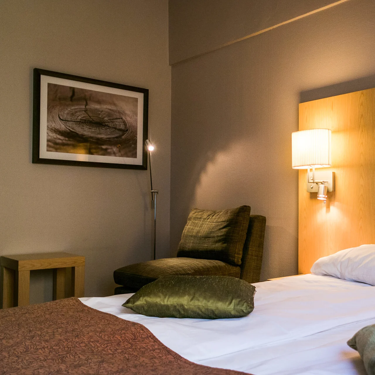 Bed with a wooden headboard, adorned with pillows and blankets, situated in a warmly-lit, cozy bedroom with an armchair and wall art. A lamp shines softly overhead.