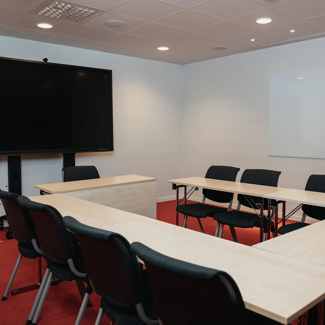 A conference room with a U-shaped arrangement of tables and chairs faces a large screen. A whiteboard is mounted on the wall in a carpeted room.
