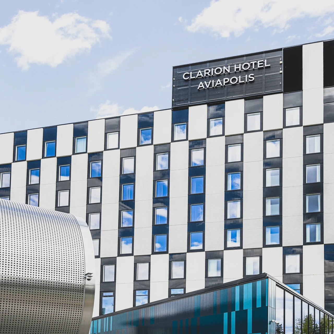 A modern, black-and-white checkered hotel building stands under a blue sky, with a cylindrical metallic structure in the foreground. Signage reads: "CLARION HOTEL AVIAPOLIS".