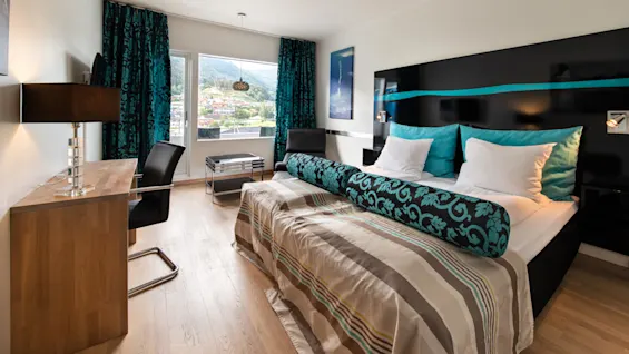 En snyggt bäddad säng med turkosa och vita kuddar står i ett modernt hotellrum. Ett skrivbord, stol och fönster med bergsutsikt kompletterar den stilrena inredningen.