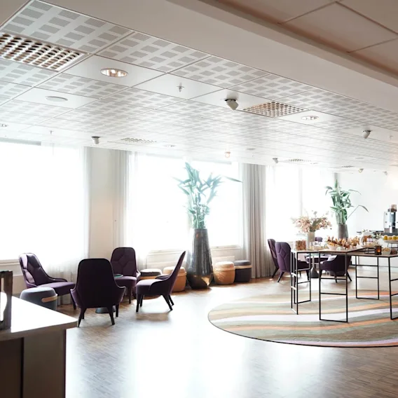 En loungeområde med lila stolar omger ett bord med bakverk och drycker. Stora krukväxter och kaffemaskiner förstärker den moderna, välbelysta miljön.