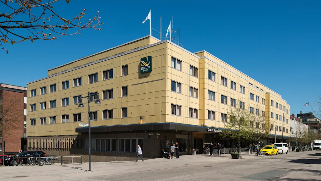 A large, yellow building labeled "Quality Hotel" with multiple windows stands on a street corner. Pedestrians walk by and a bright yellow taxi is parked nearby, under a clear sky.