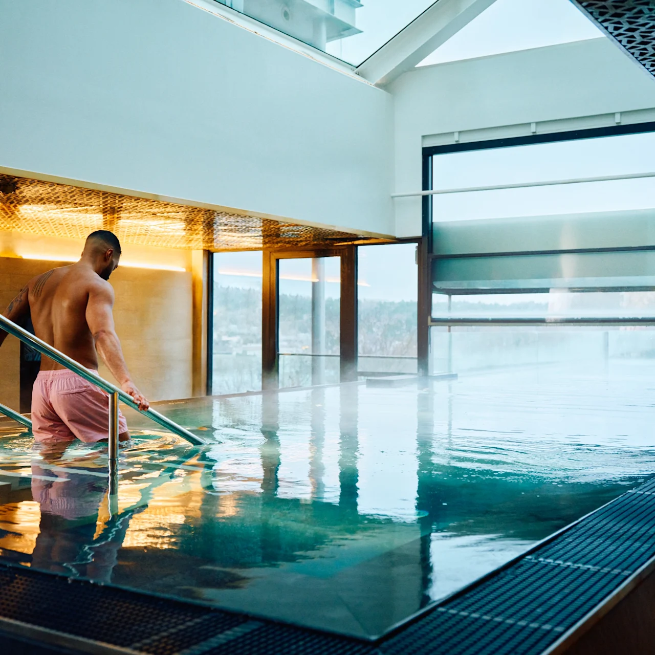 En person i rosa shorts går nerför metalltrappor i en inomhuspool, omgiven av stora fönster och modern arkitektur.