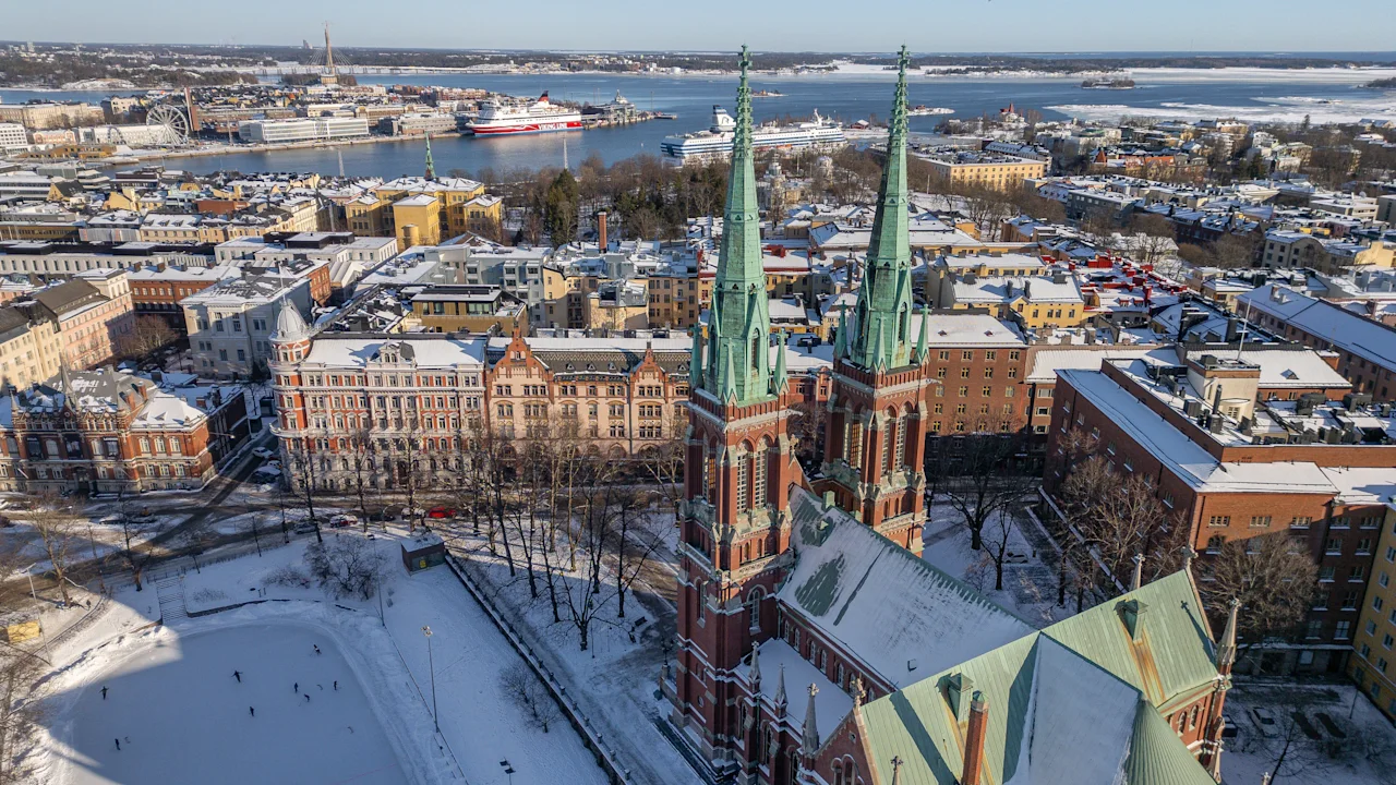 Lumipeitteinen kaupunkimaisema, jossa on suuri kirkko Helsingissä, ihmisiä luistelemassa jäätyneellä järvellä ja satama laivoineen.