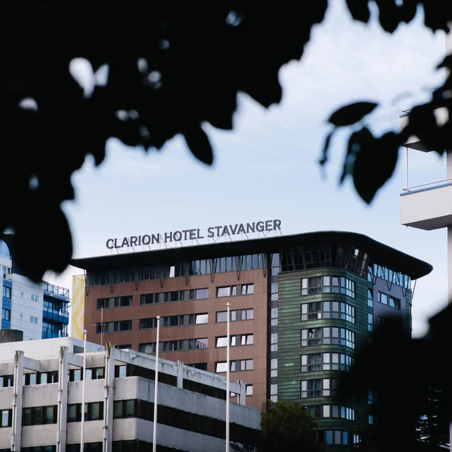 A hotel building labeled "Clarion Hotel Stavanger" stands prominently against a blue sky, surrounded by other urban structures and partially framed by out-of-focus foliage in the foreground.