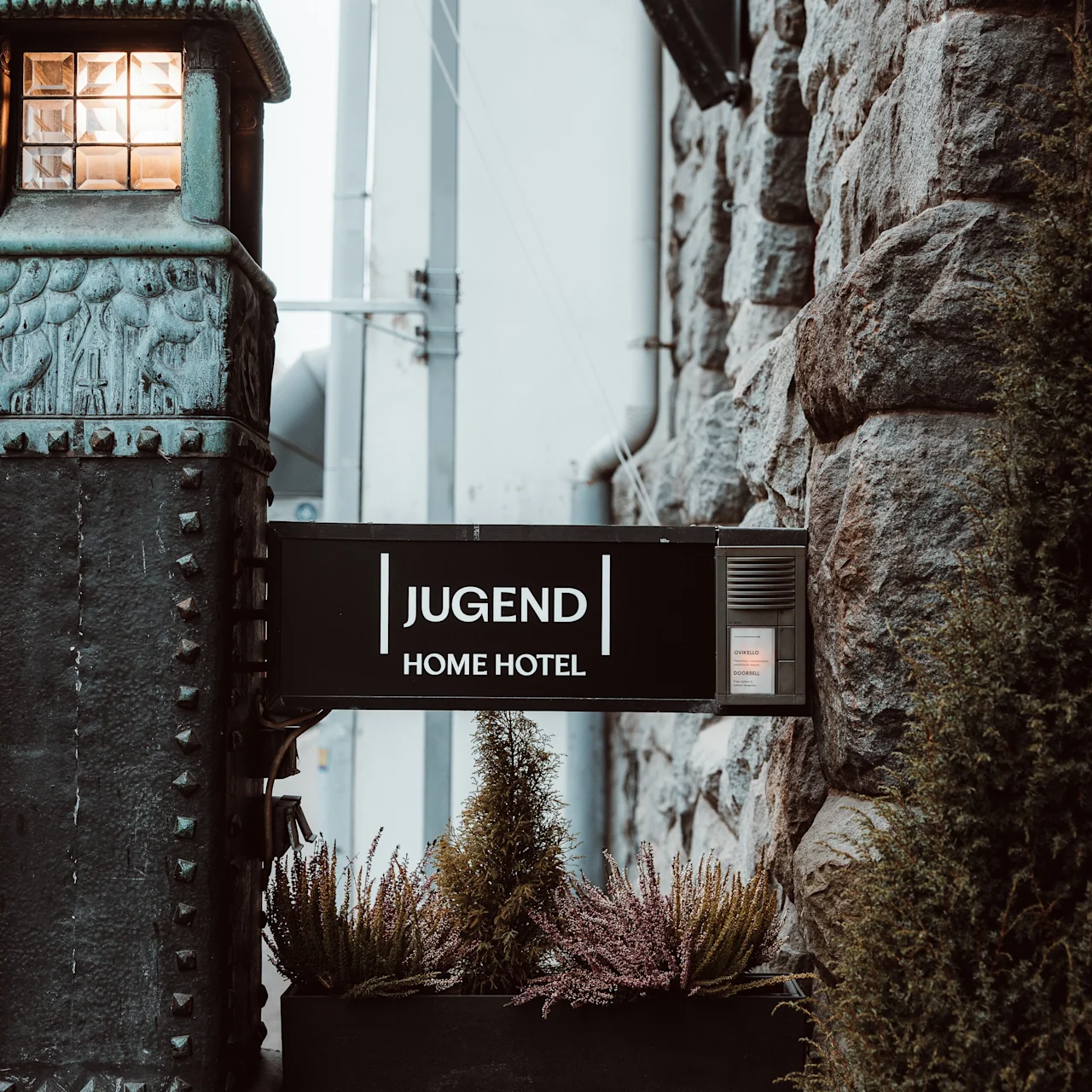 Et hotelskilt med teksten "JUGEND HOME HOTEL" monteret på en stenmur. En oplyst dekorativ lampe og planter omgiver skiltet.