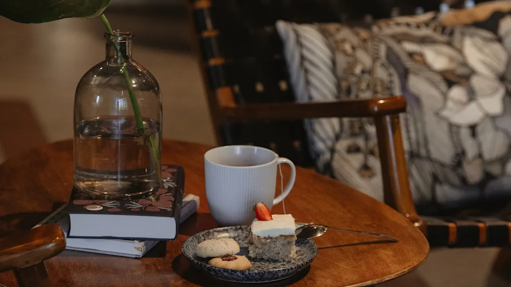 A cozy corner at Home Hotel Uppsala with a wooden table holding books, a cup of tea, and a plate of cake and cookies.