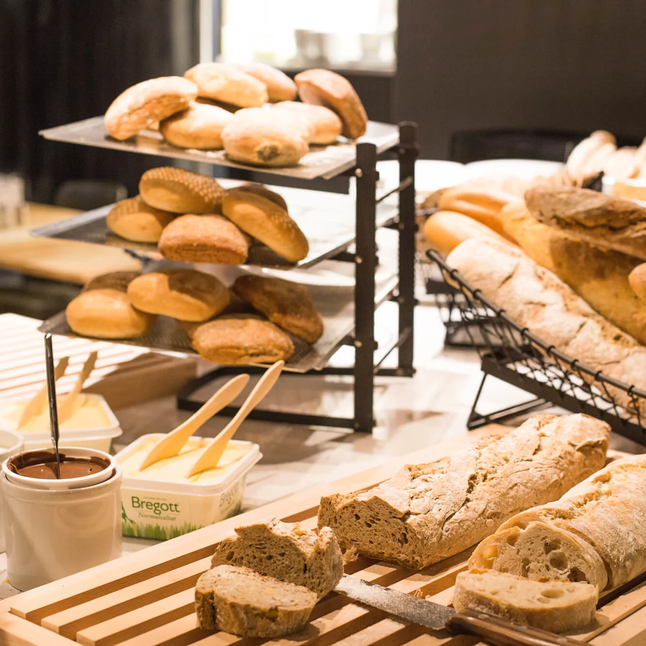 Assorted breads displayed on shelves and a wooden tray, with accompaniments like butter and spreads in a restaurant buffet setting.