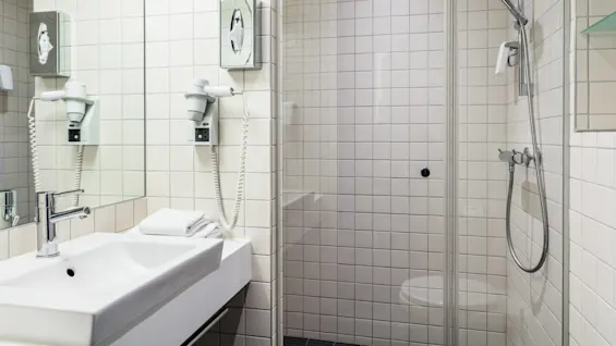 Sink with chrome faucet, towel stack beside, and wall-mounted hairdryer in a tiled bathroom featuring a glass shower enclosure and visible showerhead. 