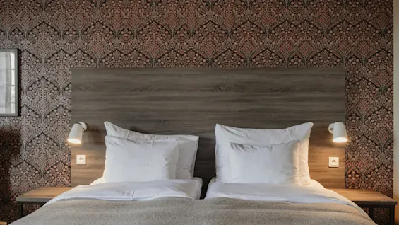 Ett mysigt hotellsovrum med en säng, sänggavel i trä och mönstrad tapet. Vägglampor ger mjuk belysning.