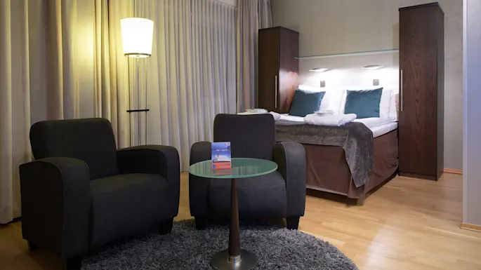 A well-lit modern hotel room with two armchairs, a glass table, and a bed in the background, setting a cozy ambiance.